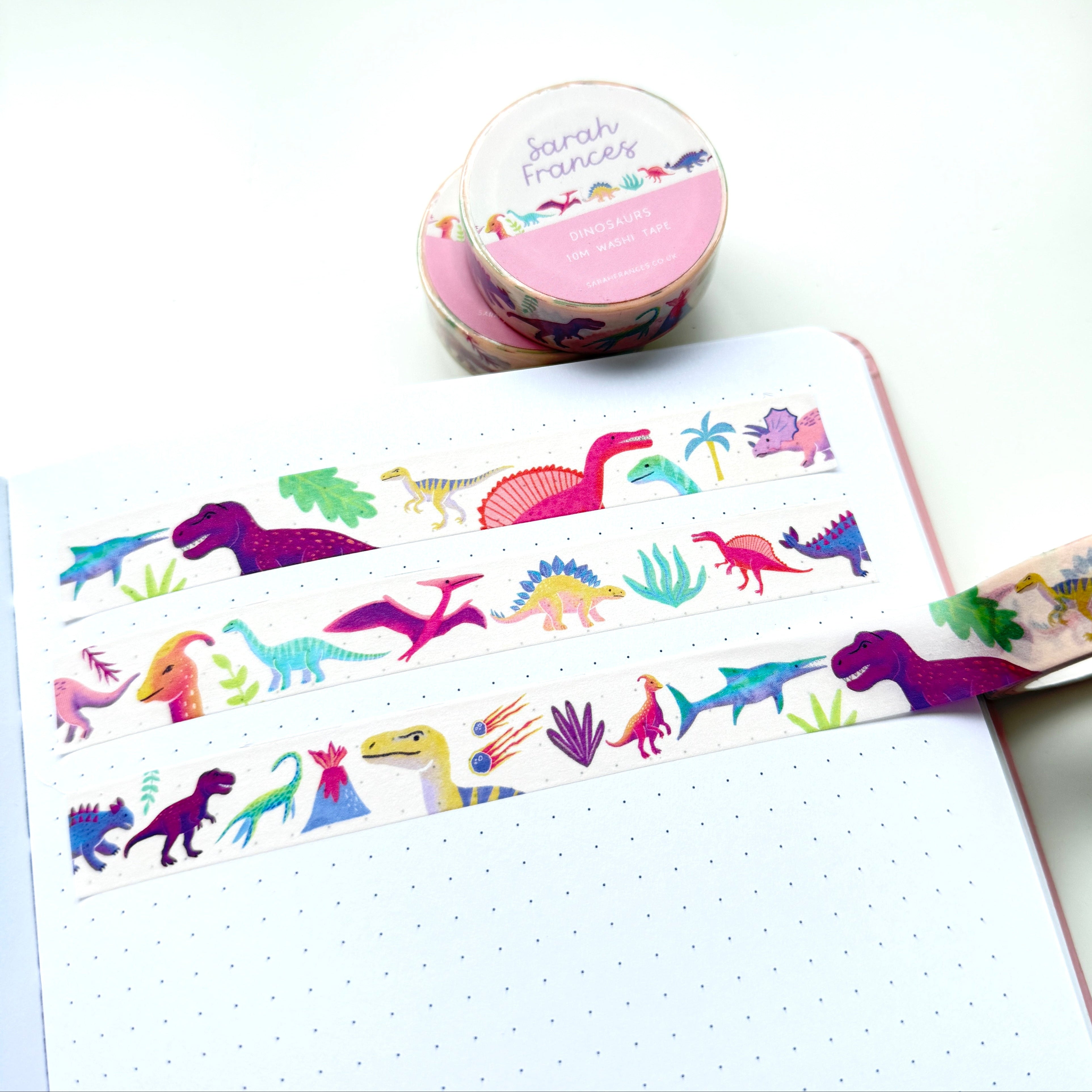 A roll of dinosaur-patterned washi tape is unrolled across an open dotted notebook, with the tape roll and a second roll visible in the background on a clean white surface.