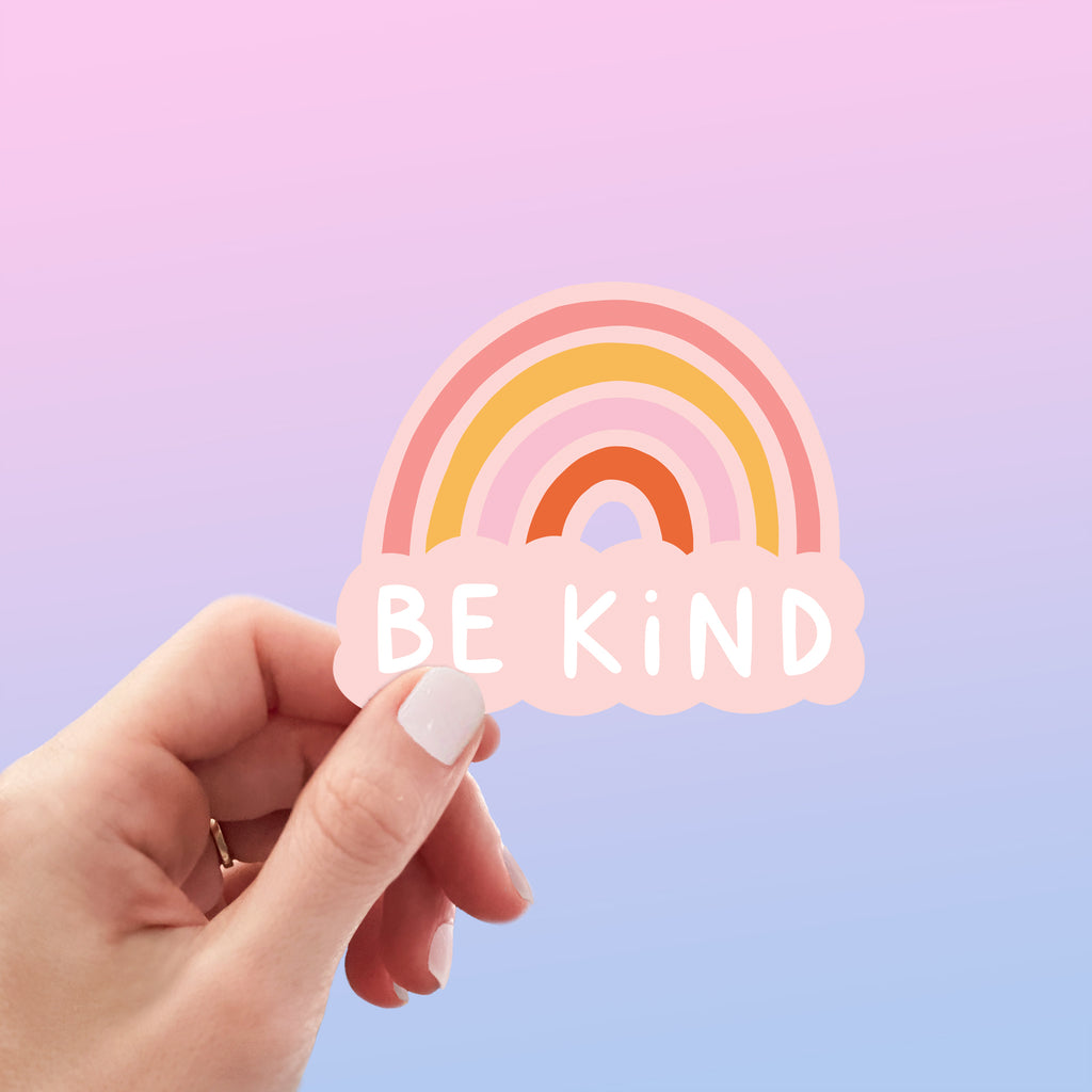 A hand holding a vinyl sticker which says Be Kind underneath a colourful rainbow.