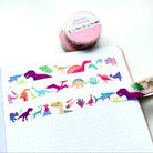 A roll of dinosaur-patterned washi tape is unrolled across an open dotted notebook, with the tape roll and a second roll visible in the background on a clean white surface.